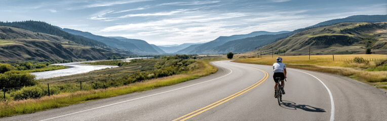 Fototapeta premium Cyclist navigates a winding road surrounded by mountains and a river under a clear blue sky during the day