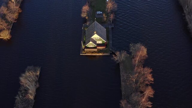 Aerial view of a quaint house surrounded by dark water channels and narrow strips of land dotted with trees, Scheendijk, Utrecht, Netherlands.