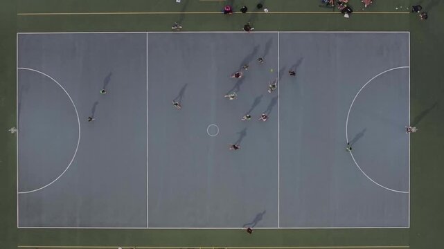 Aerial view of a netball court where players in contrasting uniforms engage in a game, their shadows stretching across the blue surface, Madeley, Western Australia, Australia.