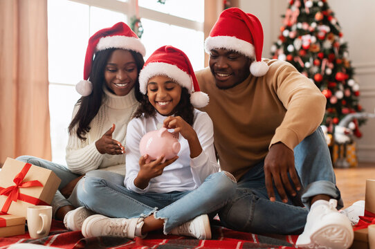 Happy African American Girl Holding Piggybank Sitting On Floor At Home, Saving Money For Future, Adding Money Putting Coin To Piggy Bank, Parents Teaching Daughter Financial Literacy. Xmas Present.
