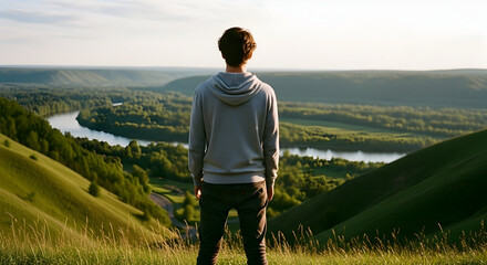 Young man standing on lush green hillside overlooking a scenic river valley with rolling hills and a bright sky in the distance capturing peaceful outdoor nature moments for travel and adventure theme