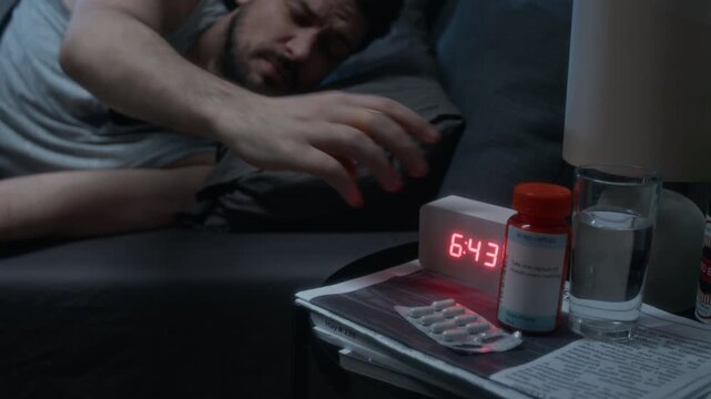 Close up of digital alarm clock with 6am on display at bedside table and restless man reaching out suffering from insomnia