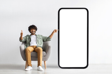 Happy handsome curly young indian man relaxing in armchair at home, pointing at huge modern mobile phone with white empty screen, recommending mobile app, panorama with copy space, mockup