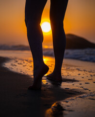 Barefoot person walking on wet sand at beach during sunrise. Close up of legs with ocean waves and glowing sun. Seaside walk and relaxation concept. 