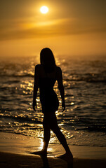 Silhouette of woman walking along beach at sunrise with glowing sun and ocean waves. Seaside walk and summer vacation concept
