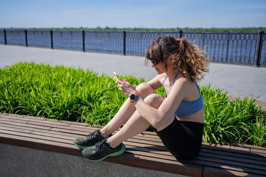 Young sportswoman in stylish sportswear and sunglasses, sitting on a bench in a city park, using her smartphone to check messages and browse social media after an outdoor workout