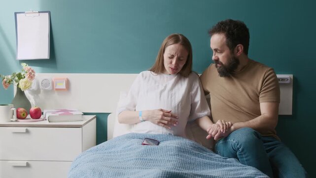 Medium full shot of pregnant young woman sitting in hospital bed with caring husband, viewing baby ultrasound images on smartphone, wife feeling painful contractions, grimacing, gripping man by hand
