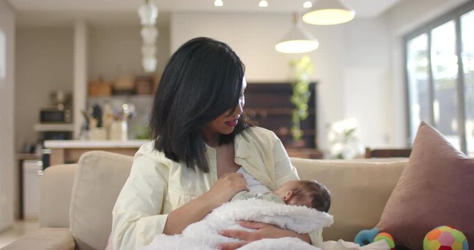 Mother lifting baby to chest for feeding while stroking head, adjusting grip on sofa near window