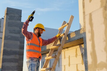 Construction worker using cordless drill on wooden ladder at building site