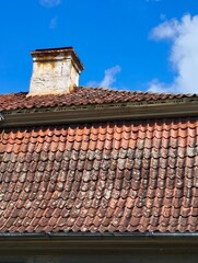 Weathered red clay tile roof with aged chimney under bright blue sky. Lichen-covered tiles reveal the charm of historic rural architecture.