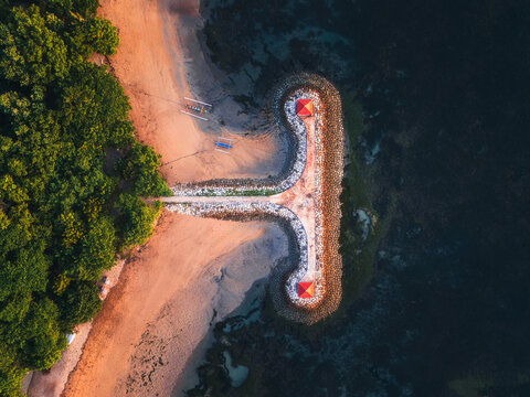 Aerial view of a T-shaped pier jutting into the dark, inviting ocean, contrasting with the golden sands and lush greenery, Sanur, Denpasar, Bali, Indonesia.