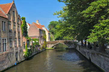 Naklejka premium A tranquil canal scene in Bruges, Belgium, featuring historic buildings, lush trees, and a classic stone bridge.