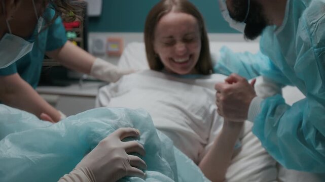 Medium shot of exhausted pregnant woman breathing heavily during final stage of labor, screaming in intense pain, pushing while giving birth to baby, midwifes assisting, loving husband holding hand
