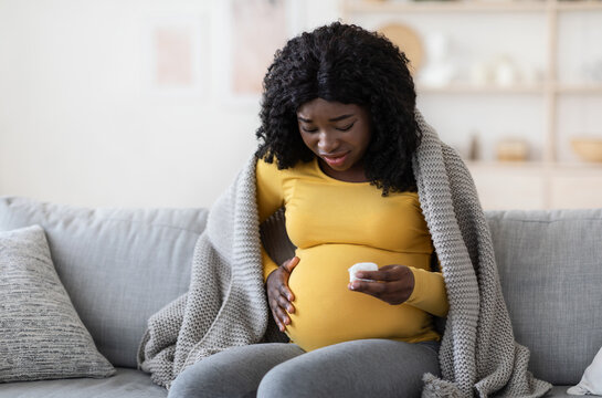 Pregnant black woman covered in blanket feeling sick, sitting on couch at home, holding napkin, worrying about her baby, empty space. Expecting african american lady having cold or flue