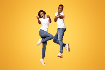 Cheerful african american young couple indicating at camera and screaming, jumping up over orange studio background. Happy black man and woman celebrating success, full length photo