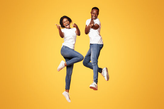 Cheerful african american young couple indicating at camera and screaming, jumping up over orange studio background. Happy black man and woman celebrating success, full length photo - Powered by Adobe