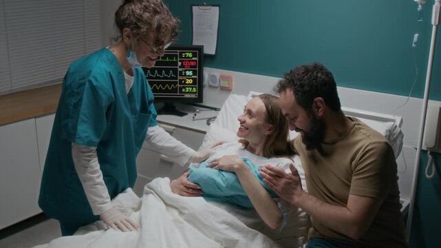 Medium shot of happy Caucasian husband and wife cradling newborn baby in delivery room in maternity hospital after labor, admiring face, while chatting to delighted midwife patting woman on shoulder