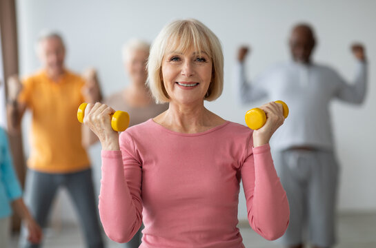 Selective focus on cheerful senior lady posing with fitness tools, multiracial group of healthy elderly people in sportswear doing strength building workout with dumbbells at gym