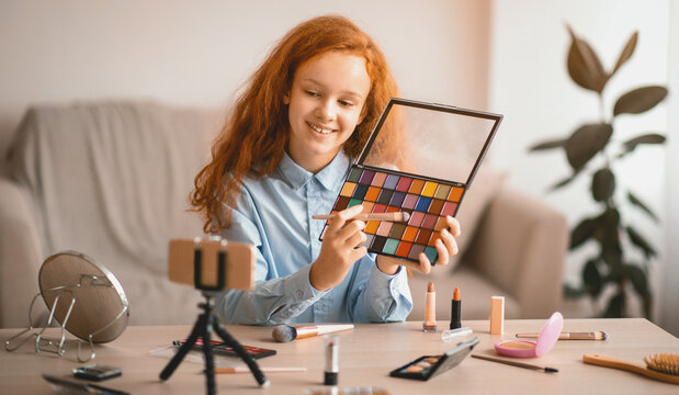 Beauty Blog. Young beautiful teen doing makeup, recording cosmetics product review, showing her eyeshadow palette to camera. Girl talking in front of phone on tripod, filming video for her channel - Powered by Adobe