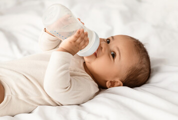 Feeding And Babycare. Closeup portrait of adorable smiling black baby holding bottle and drinking water, wearing bodysuit and resting on the white bedsheets in bedroom, lying on the back