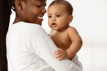 Closeup Portrait Of Cheerful African Mom Posing With Adorable Baby Boy Carrying Cute Little Son In Arms Standing In Bedroom Indoors. Motherhood Moments, Child Care And Maternity Leave, Selective Focus