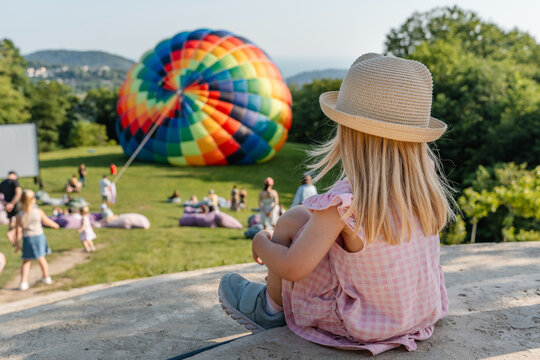 A little child girl sits on the grass and admires how a big hot air balloon is launched into the air