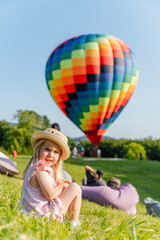 A little child girl sits on the grass and admires how a big hot air balloon is launched into the air © KseniaJoyg