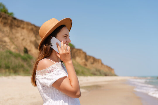 A smiling young woman in a white dress and straw hat stands on a sandy beach, talking on a smartphone, with cliffs and the ocean in the background.