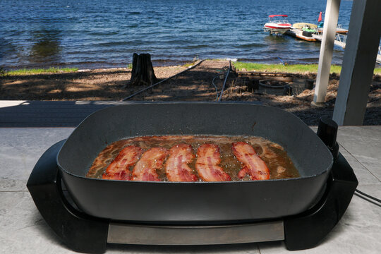 Frying hot greasy bacon for breakfast meal in oil next to a lake on a skillet grill. 