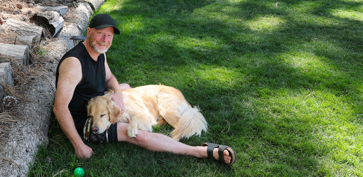 Middle aged man sitting on green grass with his golden retriever dog sleeping in his lap. 