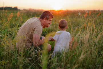 A father kneels in a grassy field, smiling at his young child who stands facing the beautiful...