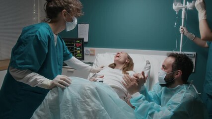 Full dolly-in shot of pregnant Caucasian woman giving birth to baby in hospital, breathing and screaming in labor, husband holding hand, midwifes setting up intravenous drip and monitoring vitals