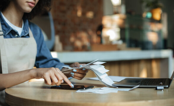 Reporting and accounting during covid-19 quarantine. Busy african american lady owner in apron works with financial bills and calculates profit and debt in modern cafe, empty space, cropped