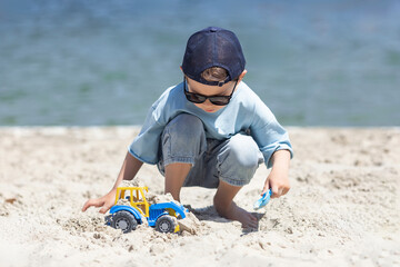 Barefoot happy little boy playing with toy car on the beach on summer holidays. Child has fun with...