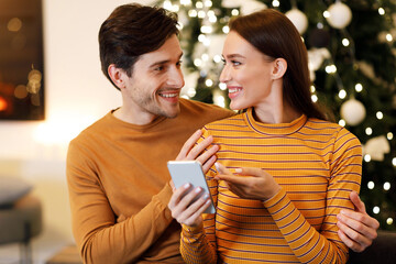 People And Technology Concept. Cheerful millennial couple sitting on couch at home using cell phone...
