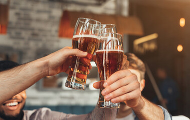 Male friends clinking beer glasses, sitting in bar, closeup