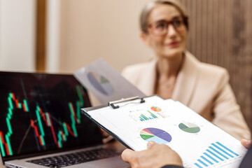 Business professional presenting financial data on clipboard to colleague, with laptop displaying stock market graphs, showcasing teamwork and analysis in a modern office environment