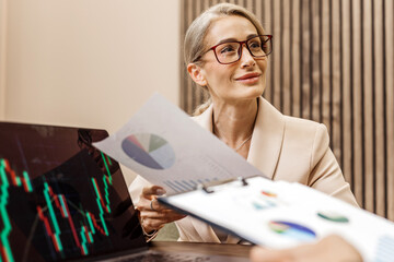 Businesswoman in beige suit, analyzing financial charts on laptop while discussing strategic planning with colleague, showcasing teamwork and professional growth in modern office environment