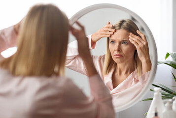 Naklejka premium Middle Aged Woman Looking In Mirror And Touching Wrinkles On Her Face, Upset Beautiful Mature Female Examining Fine Lines On Forehead, Suffering Skin Aging, Selective Focus On Reflection, Closeup
