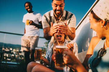 Friends enjoying drinks at a rooftop party, man surprising woman covering her eyes