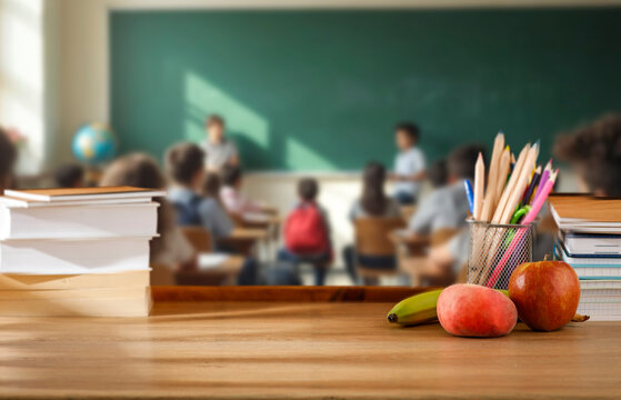 School desk with books and supplies, blurred students and teacher in classroom background. School-related atmosphere of learning, back-to-school excitement, and education in progress.