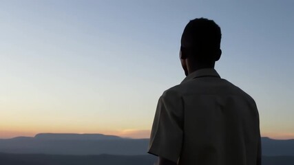 A young Black man watches the beautiful sunrise over the mountains in a peaceful and contemplative mood