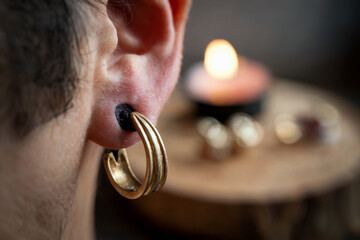 Close-up of a stretched male ear with a black plug and golden ring inserted. Warm candlelight in the background adds a cozy atmosphere.