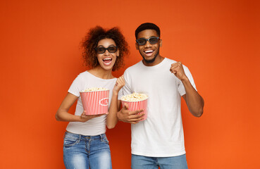 Young laughing couple in 3d glasses watching movie film on date, holding buckets of popcorn, orange studio background © Prostock-studio