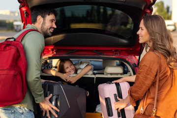 Parents packing luggage in trunk of car with kid girl sitting on backseat and looking at mother and father, family going on vacation
