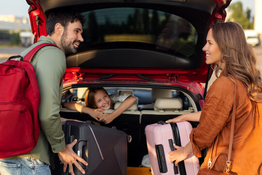 Parents packing luggage in trunk of car with kid girl sitting on backseat and looking at mother and father, family going on vacation