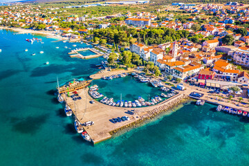 Drone image of Sveti Filip i Jakov showing coastal town layout, marina with boats, clear Adriatic sea, red rooftops, waterfront promenade and surrounding Mediterranean greenery