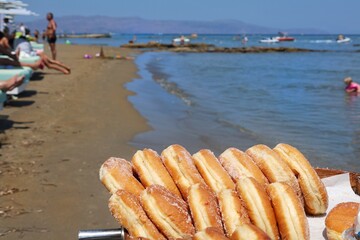 Sugar-coated donuts on a tray by the seaside at a busy beach in Crete, Greece. Popular summer street food in a holiday setting with sunbathers and boats in the background. July 2025.