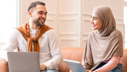 Young Muslim Couple Using Laptop And Digital Tablet Sitting On Couch And Browsing Internet Together...