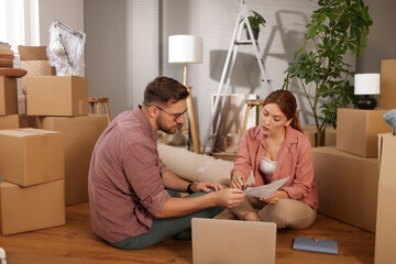 Two individuals sit on the floor, surrounded by unpacked boxes, discussing plans for their new home...